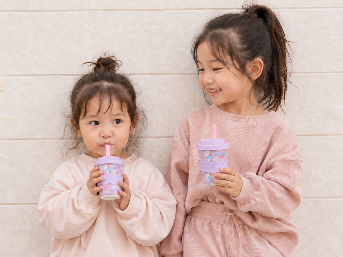 Two young girls holding Chino Club Mermaid Unicorn reusable cups, with the younger child sipping through a straw.